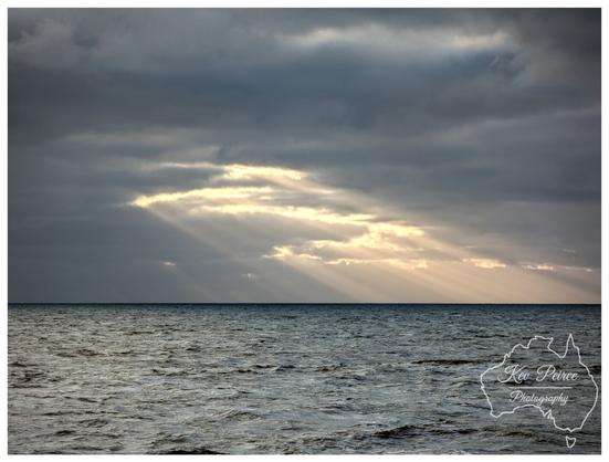 A dramatic landscape photograph taken on the Bunbury coast, showing a vast, dark, textured ocean filling the lower two thirds of the frame. Above, the sky is heavy with dark grey, overcast clouds. A central break in the clouds allows bright, warm sunlight to stream down in distinct rays (crepuscular rays) onto the water and horizon.
