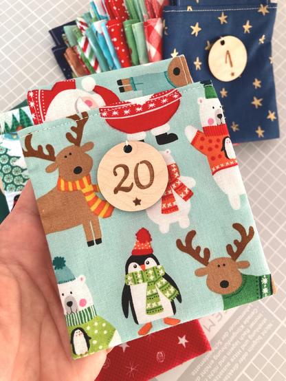 In der Mitte des Bildes ein Adventskalendersäckchen aus einem bunt bedruckten Stoff mit Pinguin, Eisbär und Rentier, die Schals, Mützen und Pullover tragen. Auf der Vorderseite ist ein runder Holzanhänger mit der Zahl 20 angenäht. Im Hintergrund liegen aufgefächert weitere Adventskalendersäckchen aus anderen Stoffen.