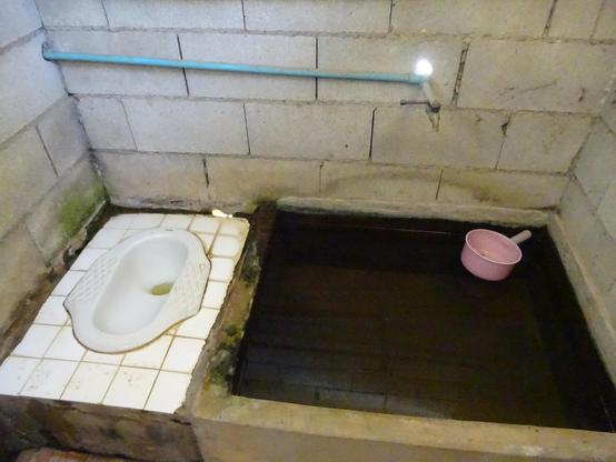 Ein einfacher Raum mit grauen Betonblockwänden. Links befindet sich eine weiße Hocktoilette auf weißen Fliesen, die teilweise verschmutzt sind. Rechts daneben ist ein rechteckiges Wasserbecken mit dunklem Wasser, in dem ein rosa Kunststoffschöpflöffel liegt. Über dem Becken verläuft eine blaue Wasserleitung mit einem Wasserhahn, aus dem Wasser tropft.