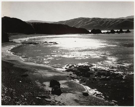 The black and white photograph captures a tranquil coastal scene, likely taken during low tide. It shows an expansive view of the shoreline leading into calm waters with scattered rocks protruding above water level. The foreground features darkened sand mixed with pools of shallow water reflecting light patches from the sun's rays that pierce through what appears to be either early morning or late afternoon skies.

A line of large, rugged cliffs dominates one side of the beach, creating a dramatic silhouette against an overcast sky while their shadows stretch across the sandy beach. In contrast, on the far right edge of the photo, smaller rocky formations break the water's surface in the distance with no land immediately visible behind them. The absence of any significant human presence or man-made structures lends to the scene’s natural and untouched quality.

The photograph exhibits a rich play of light and shadow which adds depth and texture throughout the landscape; from the fine grains on the sand, dark recesses within crevices between rocks near shore, as well as variations in brightness across water patches. This interplay enhances the overall atmosphere of serene isolation characteristic of such coastal environments.

There is no discernible movement or action captured in this still shot, suggesting a moment frozen in time which invites contemplation on nature's quiet beauty and power.