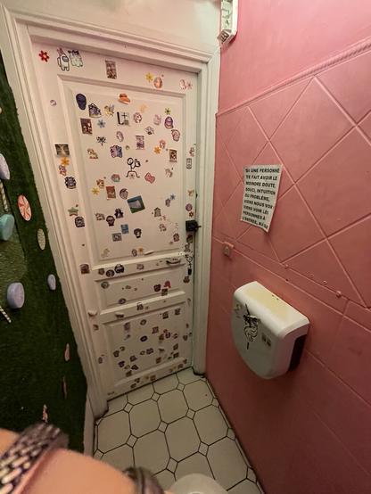 A cozy tiny room with a framed door partly covered with tiny LGBT and cute stickers. One wall to the right is pink, the other to the left is covered with fake green grass.