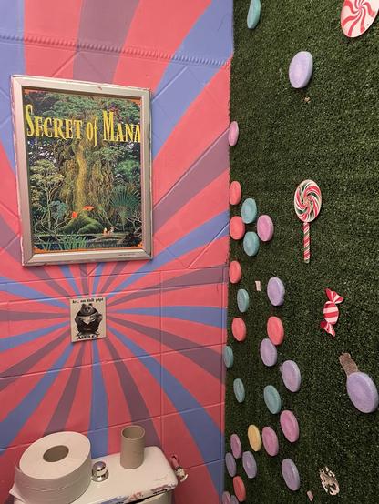 The fake green grass is plastered with lollipops, candies and various round colored ornaments. The last wall is painted with a bright pink, blue and lavender pattern and features a Secret of Mana poster.