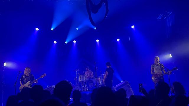 Les membres du groupe de punk Pogo Car Crash Control sur scène avec de gauche à droite la bassiste, le batteur au fond, le chanteur principal et le guitariste. L’atmosphère est bleutée. En hauteur, accroché au plafond, il y a un grappin de machine à grappin.