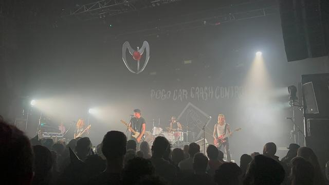 Les membres du groupe de punk Pogo Car Crash Control sur scène avec de gauche à droite la bassiste, le chanteur principal, le batteur au fond et le guitariste. L’atmosphère est bleutée. En hauteur, accroché au plafond, il y a un grappin de machine à grappin.