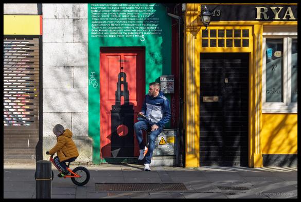 Vibrant street art photograph showing a child on a balance bike wearing a gold jacket, an adult sitting on an electricity box in front of street murals with a turquoise wall listing local Cork businesses in white text, a red doorway with a black bottle silhouette, and a bright yellow storefront numbered 43 on North Main Street, Cork.