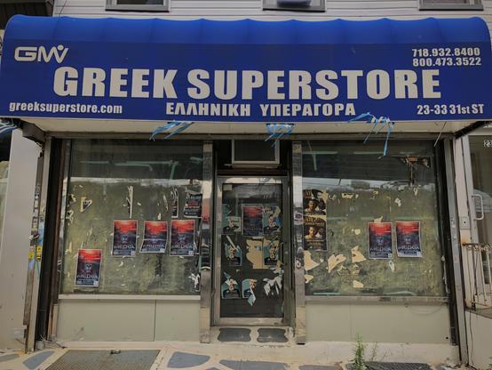 A blue and white banner reads “Greek Superstore” in English and Greek. The store is shuttered with the front plastered with stickers and ads.