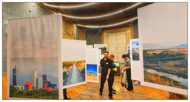 Un interno a una galleria d'arte con un pavimento di legno e pareti bianche. Diverse persone sono visibili mentre guardano le immagini esposte. La stanza è illuminata da luci a soffitto. Ci sono una serie di immagini incorniciate appese alle pareti, che variano in dimensioni e colore. C'è un'immagine di grattacieli, una cascata e un paesaggio con un fiume.