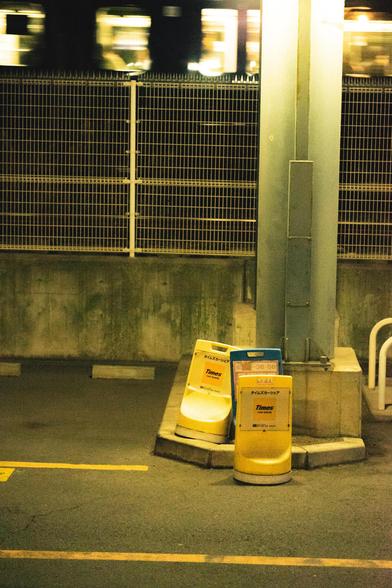 Dimly lit parking area with three yellow and blue traffic barriers labeled "Times" next to a large metal pole. A high fence runs along the background with a blurred train passing by above. The ground has yellow painted lines indicating parking spaces, and a concrete wall is visible behind the fence.