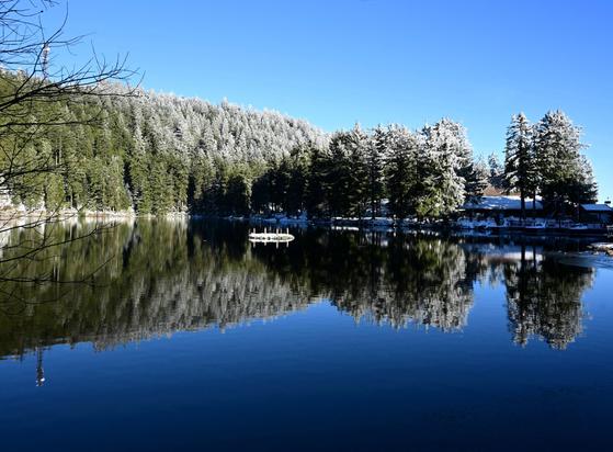 Blauer See umrandet von teilweise mit Schnee bedeckten Bäume, die sich im Wasser spiegeln. Inmitten des Sees befindet sich ein Adventskranz, der sich ebenfalls spiegelt.