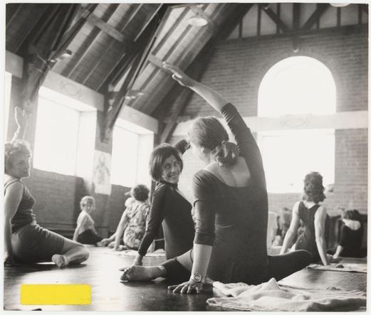 The black and white photograph depicts an indoor scene focused on a group of individuals engaged in what appears to be physical exercise, possibly yoga or stretching. The central figure is seated with legs extended along the floor while extending one arm upwards, demonstrating flexibility and balance. This person wears dark clothing suitable for movement. To their right sits another individual facing away from the camera, seemingly observing the activity. In front of them are several people in various states of relaxation; some sit on cushions or directly on the floor with legs extended horizontally.

The room features exposed wooden beams supporting a vaulted ceiling and large windows that allow natural light to enter, creating bright areas where sunlight streams through. A distinctive arched window provides additional illumination near the far wall. The group is spread across the expanse of what seems like an old hall or studio space with a high ceiling.

The image captures a candid moment among participants who display focused and serene expressions, suggesting a shared activity that promotes wellness or mindfulness. There are no visible texts within the photograph to indicate location, event specifics, or any identifiable information about the individuals present.