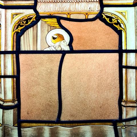 A painted glass church window made of four panels which come together to show a complete picture.  The first shows part of a window shows a bearded head and the tip of a golden wing. The other three panels have been replaced by plain glass.