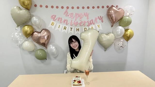 A woman smiling holding a large 1 balloon with a birthday cake on the table with happy anniversary birthday banner hang up on the wall with an assortment of balloons.