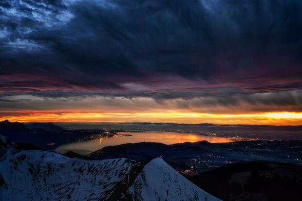 Diese Weitwinkelaufnahme zeigt eine majestätische Alpenlandschaft bei Sonnenuntergang, aufgenommen von einem schneebedeckten Berggipfel. Im Vordergrund erstrecken sich verschneite Hänge, während im Mittelgrund der Genfersee von den Lichtern der umliegenden Städte beleuchtet wird, die sich auf der Wasseroberfläche spiegeln. Der Himmel ist von dramatischen Wolkenformationen geprägt, die in leuchtenden Orange-, Gelb- und Rottönen erstrahlen und einen starken Kontrast zu den dunkleren Blautönen bilden.