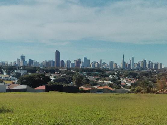 Panorama em um dia parcialmente nublado de uma cidade com muitos edifícios no horizonte, vistos a alguns quilômetros a partir de um terreno com grama verde. Entre arranha-céus no horizonte, há uma alta edificação em formato diferente, cônico, que é a Catedral Nossa Senhora da Glória. A área intermediária parece predominantemente residencial, com edificações baixas entre muitas árvores.