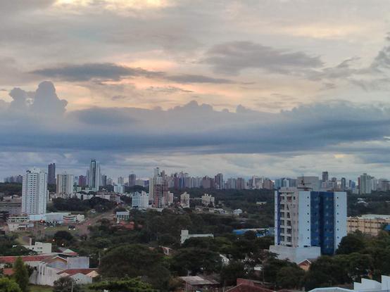 Panorama em meio a uma cidade em fim de dia nublado, a partir de uma posição um pouco elevada do solo, contemplando o horizonte com muitos edifícios e, até lá, zonas mistas com muitíssima árborização verde.