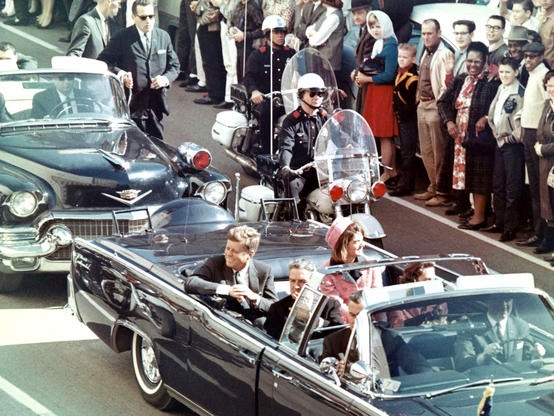 Photo du président Kennedy, souriant dans sa limousine décapotable, entouré par un cortège de voitures et motos, quelques minutes avant son assassinat à Dallas en 1963.