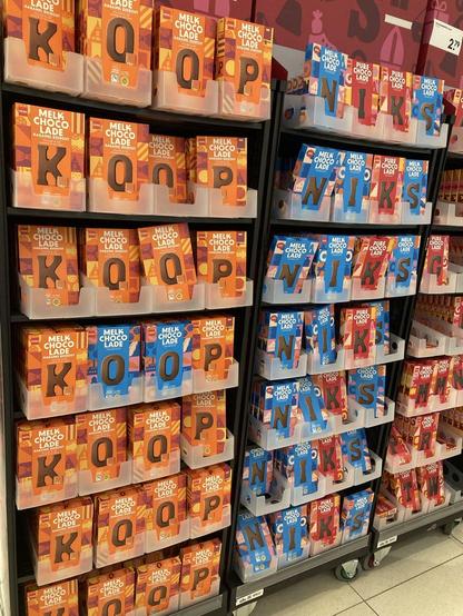 7 rijen naar benedern en 2 rekken met 4 choco letters naast elkaar
eerste rek vol met oranje verpakkingen met bruine letter, op 2 na, in blauwe verpakking
2e rek vnl blauwe verpakking op de K na, die is rood met bruine letter