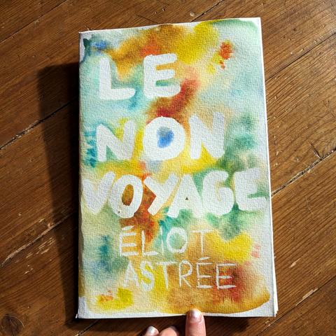 La première de couverture du fanzine posé sur un parquet. Le fanzine est peint : il y a des fonds abstraits et multicolores à la peinture à l'eau, le texte apparaît dans des zones laissées blanches. On voit le titre "le non voyage" et le non de l'auteur "éliot astrée". La couverture est dans les tons bleu clairs, vert d'eau et ocre.