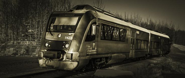 Sepia-toned image of a modern passenger train labeled 1423 moving through a snowy forest landscape. The train contrasts with the leafless trees and snow-covered ground, evoking a sense of quiet transit in winter.