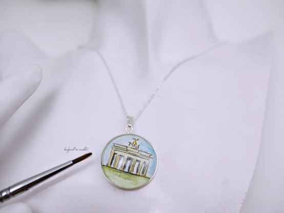 Ein winziges handgemaltes Aquarell mit 25mm-Miniatur des Berliner Brandenburger Tors. Auf rohweißem rauhem Echtbüttenpapier gemalt und unter Glas geschützt
Runde 25mm- Fassung aus 925 Sterling Silber mit passender 45cm-Ankerkette, ebenfalls aus 925 Sterling Silber , an Schaufensterpuppe fotografiert