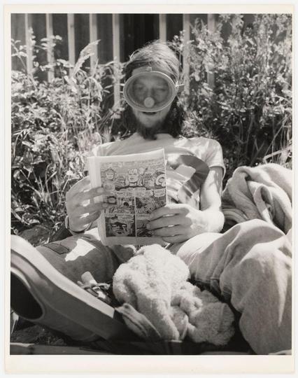 The black and white photograph depicts a person seated outdoors, surrounded by foliage. The individual is wearing a large diving mask over their face, which covers the eyes but leaves the mouth exposed for breathing or speaking purposes. They are engrossed in reading a comic book with visible panels that include characters and speech bubbles.

In front of them on the ground lie several plush toys, suggesting a casual, relaxed setting possibly indicating leisure time spent outdoors. The sunlight filters through, casting shadows across their body and creating a peaceful ambiance.

The photograph is characterized by its stark contrasts between light and shadow, contributing to a timeless quality. This image could evoke curiosity about the person's circumstances or story, hinting at themes of isolation, creativity, or unconventional hobbies.