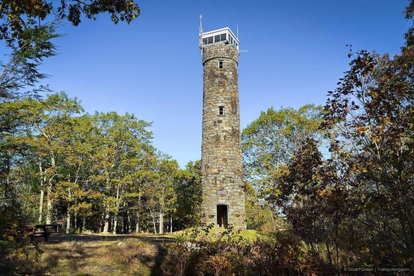 A tall, cylindrical stone tower rises from a forest clearing, built from irregularly shaped stones and topped with a small observation deck featuring a white railing and antenna. Surrounding deciduous trees display green and yellow foliage, suggesting early autumn. A picnic table sits nearby, and the clear blue sky adds to the peaceful, scenic atmosphere.