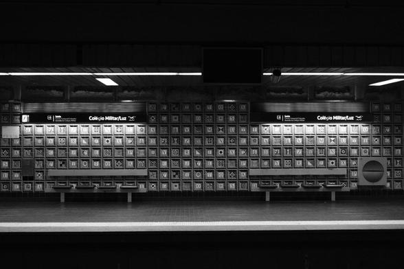 BW picture of the empty Colégio Militar/ Luz Subway station, with two empty benches and above them the sign identifying the Station by Name.
