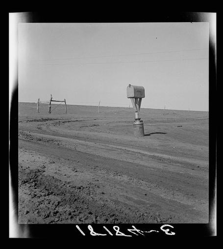 A black and white photograph depicting a rural landscape with minimal elements. In the center, there's an elevated mailbox on four wooden posts in what appears to be arid terrain, indicative of a Dust Bowl region or similar dry climate area. The ground is uneven and covered with small clumps of earth or dirt. To the left side stands a dilapidated fence structure made from simple poles and horizontal elements, suggesting a once maintained boundary now succumbing to neglect. Above, an overcast sky stretches across without any visible clouds, creating a somber mood in this desolate setting. The overall scene reflects isolation and abandonment common during historical periods of drought or economic hardship such as the Great Depression era's Dust Bowl phenomenon in Texas.