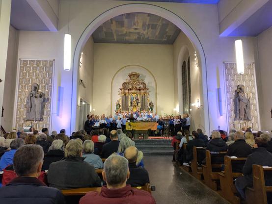 Blick in den Altarraum der katholischen Kirche. Im Vordergurnd sieht man Publikum in den Kirchenbänken, in der Mitte den ca. 40 Personen umfassenden Chor, der im Alltarraum steht. Alles ist hell und freundlich.
