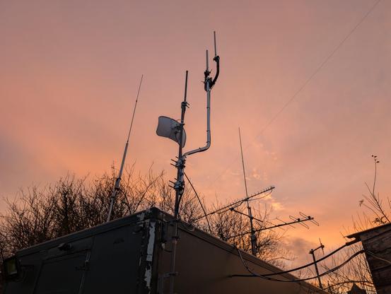 Multiple antennas are visible in the foreground mounted to a green cabin. The sky is cloudy and pinky-orange, caused by the sunset.