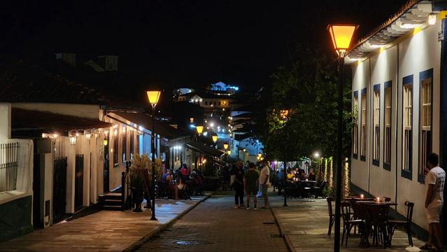 Uma rua movimentada à noite, iluminada por postes e luminárias. Edifícios antigos ladeiam a rua, com mesas e cadeiras do lado de fora de estabelecimentos, um ambiente de bar ou restaurante. Pessoas passeiam e se reúnem, enquanto no fundo, casas iluminadas sobem por uma colina, criando um cenário charmoso e acolhedor.