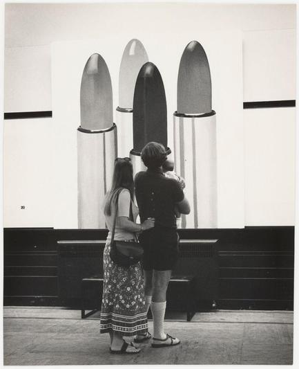 The photograph is a black and white depiction capturing two individuals engaged with an art installation. The setting appears to be indoors, possibly in a gallery or exhibition space. On display are three large objects resembling missiles or rockets that stand vertically on their fins against a plain wall backdrop. These objects vary in color: the middle one has a reflective metallic finish, while the ones at each end have darker tones with matte surfaces.

On the left side of the image, there's an individual wearing what seems to be vintage clothing—a sleeveless top and patterned skirt—with sandals on their feet. This person is turned away from the camera, looking towards or possibly touching one of the missile-like objects. To the right stands another figure in a short-sleeved dark shirt with shorts and high socks, holding an infant close against them as they gaze at the display.

The individuals' engagement suggests curiosity or contemplation regarding the art piece. The photograph has a candid quality to it, capturing a moment of interaction between people and contemporary artwork.