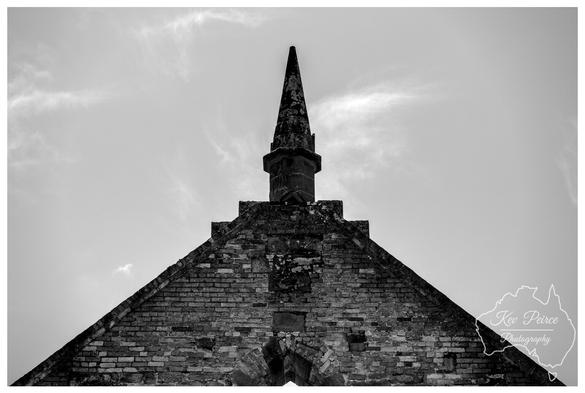 The image focuses tightly on the brick gable end and central stone spire of a historic ruined building, likely the church or main structure at Port Arthur, Tasmania.  The rough, textured brickwork fills the base of the frame, converging into a central stone spire that points dramatically upwards into a bright, cloudy sky.