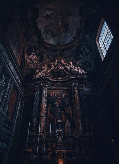 A photograph of an ornate, dimly-lit church altar and looking up to the ceiling. The main focus is an altar adorned with detailed carvings, columns, and a large dark painting depicting religious figures. Above, there are frescoes and angels are visible. The background includes a window to the left allowing limited natural light. The overall colour palette is dark with muted gold accents.