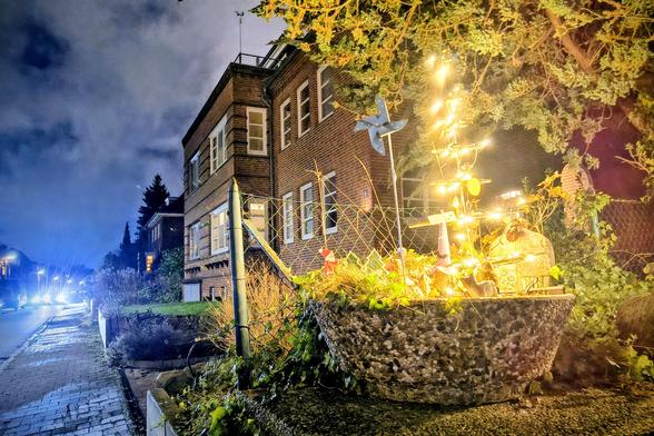 Ein weihnachtliche dekorierter Steintopf in einem Vorgarten, beleuchtet mit einer goldenen Lichterkette. Links blaues Licht des Abendhimmels und von den Scheinwerfern eines herannahenden Autos. Dazwischen ein zweigeschossiges Backsteinhaus.