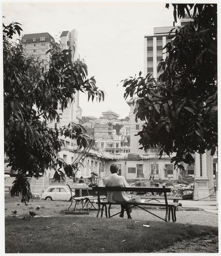 The photograph is a black and white, possibly historical snapshot capturing an individual seated on a bench in what appears to be an urban park or public garden. The person, seemingly absorbed in thought while holding reading material—likely a newspaper—is facing away from the camera towards a collection of buildings that suggest either post-war reconstruction or architectural diversity characteristic of many cityscapes.

Behind them is a mix of classical and modernist architecture; prominent among these are structures with columns and neoclassical features, which contrast sharply against more contemporary high-rise edifices. The foreground includes greenery, possibly trees or shrubs that frame the left side of the image, adding depth to this urban tableau.

The setting seems peaceful despite signs of disrepair in some buildings—a partially collapsed roof suggests recent destruction—contrasting with a sense of calm and contemplation conveyed by the person's posture on the bench. The overall atmosphere hints at resilience amidst urban decay or redevelopment. This juxtaposition between tranquil solitude and the surrounding cityscape might be indicative of themes related to personal reflection within the context of broader societal changes.

The photo is attributed to Ans Westra, as noted in a caption provided with an image reference link that directs viewers to further information about this work on Wellington - Post Offic [...]