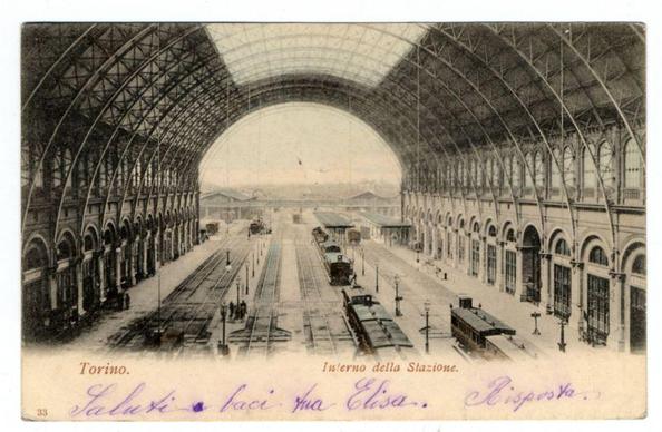The image presents a historical photograph of the interior of Turin's train station. The expansive hall is constructed with an intricate network of steel beams and glass panels, creating a spacious arched roof that allows light to filter through. In the center lies a broad railway platform flanked by tracks on both sides, leading towards what appears to be terminal buildings in the distance.

On either side of the platforms are rows of large windows or arches with rounded tops, typical of historic architecture designed for public spaces like train stations. A number of vintage tram cars and possibly early motorized trains can be seen stationed along the tracks, indicative of a bygone era. The image is tinged with sepia tones suggesting it's an old photograph.

Intricate handwritten notes in Italian are present across the bottom edge of the photo: "Salute", "Faci", "ma Elsa.", and "Bisposua" respectively, possibly representing names or phrases from a personal note added to commemorate this location. The top left corner has additional text stating "Torino." (Turin.) followed by an archaic phrase for 'Interni della Stazione', which translates as 'Interior of the Station.' A label with the number 33 is visible on the bottom right, likely indicating a cataloging or collection reference.

The photo seems to be part of a series dedicated to Italian cities and their landmarks, suggested by the consistent format used across image [...]