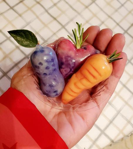 A hand holding three colorful Thai mungbean sculpted creations shaped like a purple peanut with a green leaf, a beet heart with rosemary sprigs, and an orange carrot with a green stem, all in a clear plastic dish.