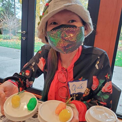 Me, wearing a sequined face mask, floral shirt, and whimsical felted wo hat, holding small dishes with decorative fruit-shaped mungbean sculptures, smiling towards the camera, with a name tag that reads "Qué" pinned on my shirt.