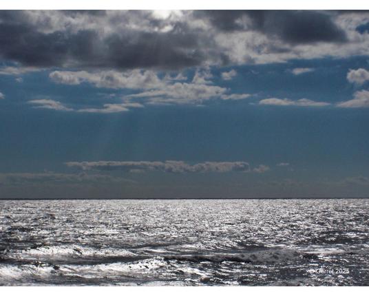 Bright sun rays peek through a cloudy sky over a shimmering ocean, creating a serene and reflective mood with a subtle, calm horizon.