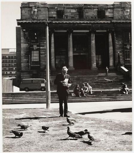 The photograph shows a man standing on what appears to be a grassy area in front of an impressive, historical building with classical architecture. The structure features prominent columns and large windows, suggesting it might be an important public or government edifice. There are several people seated on steps leading up to the entrance of this building, indicating that the space is open for visitors.

In the foreground, a group of birds—possibly pigeons—are scattered across the ground near the man's feet. The black and white nature of the image provides a stark contrast between the subjects in focus (the man and the birds) against the detailed backdrop of the architecture. A minivan is parked on the street beside the building, adding to the urban setting.

The composition captures a moment that suggests contemplation or waiting as the central figure holds what looks like papers or documents, perhaps reading them while surrounded by city life. The presence of multiple pigeons adds an element of everyday nature within this constructed environment.