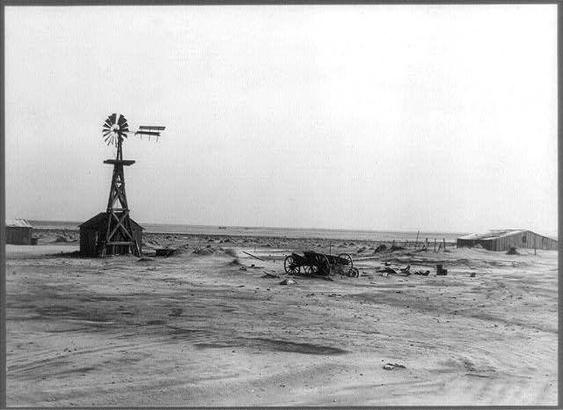 This black and white photograph captures a scene from an abandoned farm in the Coldwater District, north of Dalhart, Texas. The barren landscape is dominated by a windmill with two vanes on top, standing tall amidst scattered debris such as broken pieces of wood and machinery parts strewn across the sandy ground. To the left, there's a dilapidated shack housing another piece of equipment attached to its roof.

A weathered horse-drawn carriage sits in the foreground, adding a sense of history and abandonment to the scene. In the background, various farm buildings can be seen, including what appears to be an old barn or storage shed, further emphasizing the rural setting. The sky is overcast, casting a somber mood over this desolate landscape.

The photograph provides a glimpse into the past, showcasing how farmland and agriculture have changed through time in Texas's Coldwater District. This abandoned farm represents not only the physical remnants of agricultural practices but also hints at the social and economic conditions that may have led to its current state.
