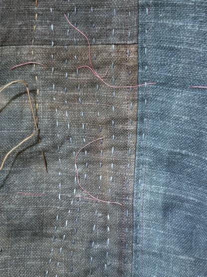 Linen fabric in various shades of indigo blue, sewn together and being quilted with densely spaced slightly wavy lines. A long needle is sticking into the fabric at the left.