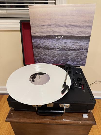 The album “Ixora” by Copeland playing on a record player. The record is a white color. The record jacket sits upright against the back. The album cover is of a wave on a beach with the sun faintly reflecting in the background. The word “Ixora” is written in white over the center.