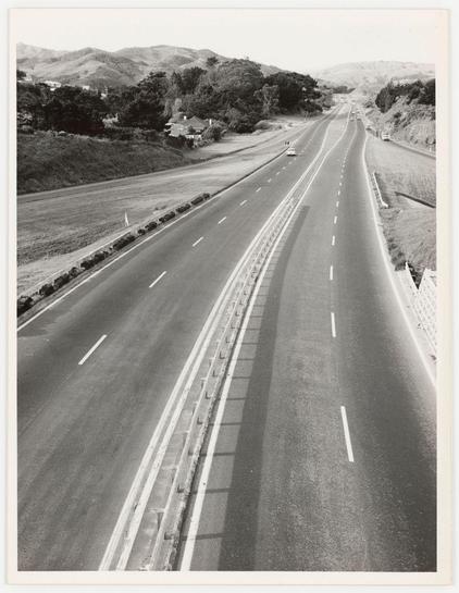 The photograph captures an aerial view of a highway, displaying two lanes in each direction. The roadway is marked with white dashed lines indicating the driving path and solid broken lines demarcating lane divisions. Alongside the road, there's a clear barrier separating the traffic from what seems to be a grassy verge on one side and elevated structures or walls on the other.

In the distance, gentle hills rise up against an overcast sky, suggesting this might be located in a region with rolling landscapes. The absence of heavy vehicular traffic suggests either low volumes during non-peak hours or possible road closure conditions not immediately visible from above.

No vehicles are captured at rest within these lanes; however, there is one small vehicle on the rightmost side, perhaps an outlier heading towards another lane or just entering the shot's field. The overall scene presents a tranquil depiction of highway infrastructure with natural surroundings in view.