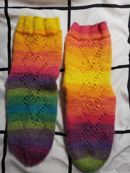 A pair of knitted socks made from rainbow yarn with a repeating heart lace pattern