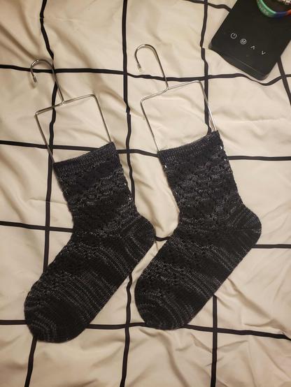 A pair of black/grey unisex knitted socks with a mottled lace pattern displayed on wire blockers