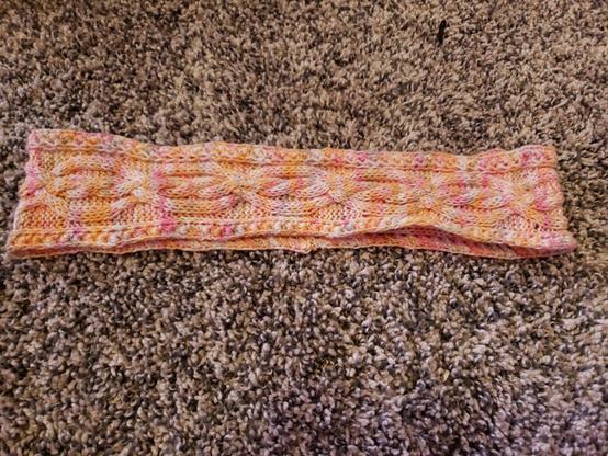 A cable knit headband laid flat made from variegated yarn in shades of pink, orange, and white--the lesbian pride flag colors