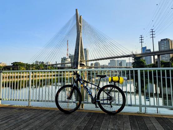 A mountain bike with a black frame and a bright yellow saddle bag is parked on a wooden boardwalk overlooking a river or canal. In the background, the Ponte Estaiada Octávio Frias de Oliveira, a distinctive cable-stayed bridge with a single pylon resembling an 'X', dominates the cityscape under a clear blue sky. High-rise buildings line the horizon, and there are power lines and a second, older bridge structure to the right.