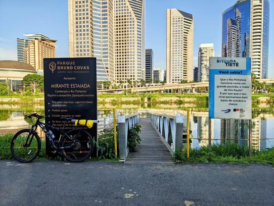 A mountain bike with a black frame and a yellow saddle bag is parked next to a black informational sign for the Parque Bruno Covas - Eixo Rio Pinheiros, specifically the MIRANTE ESTAIADA viewpoint. The sign encourages contemplation of the Pinheiros River and lists rules like prohibition of fishing, diving, or swimming. To the right, a wooden footbridge extends over the water toward a grassy bank. A second blue and white sign, possibly containing environmental information about the Pinheiros River's water level regulation, is visible on the right. In the background, a row of modern, glass-facade skyscrapers stands against a clear blue sky, with a rail line visible above the river.